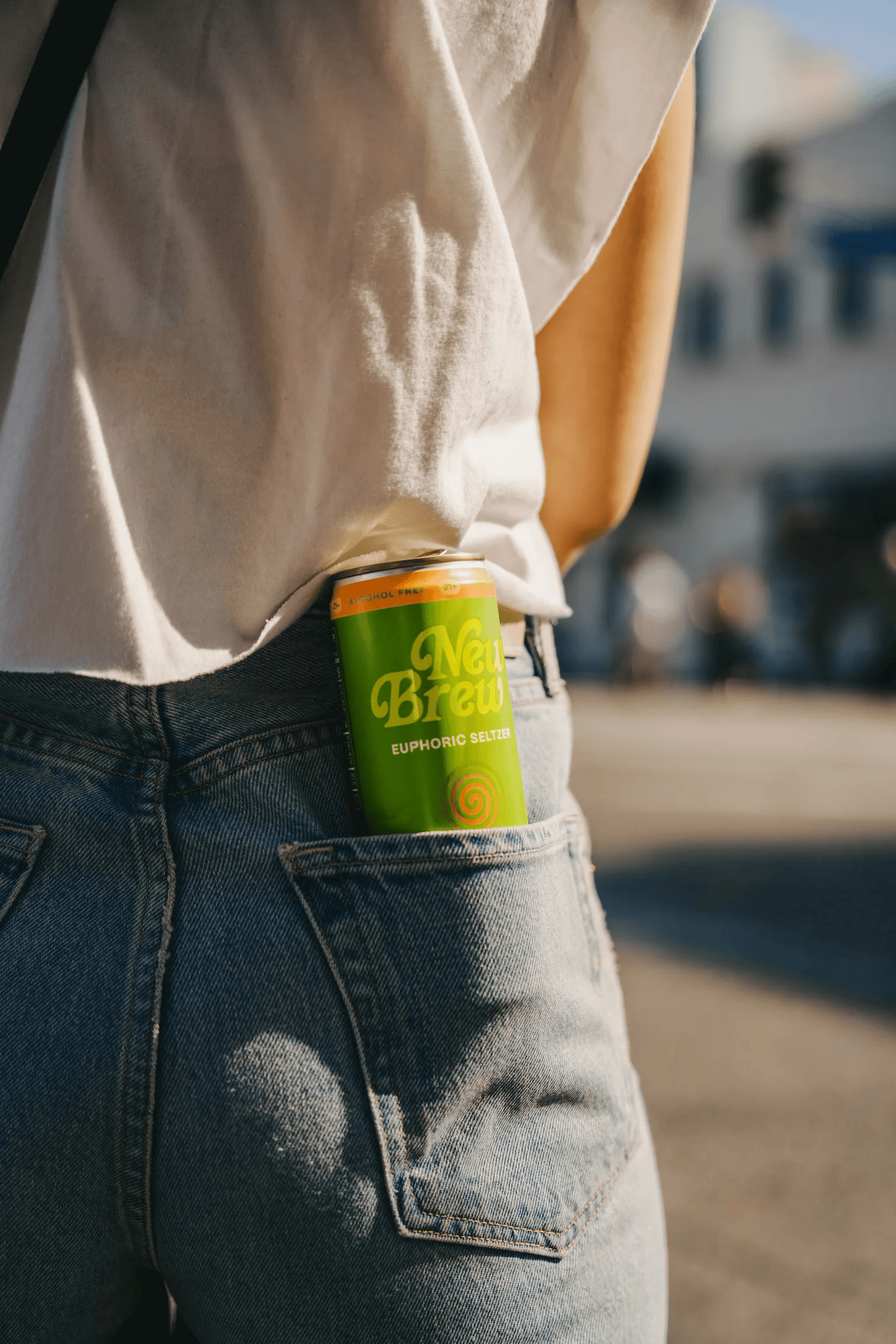 Full-color lifestyle campaign photograph of a canned beverage tucked into a denim back pocket on a sunny street
