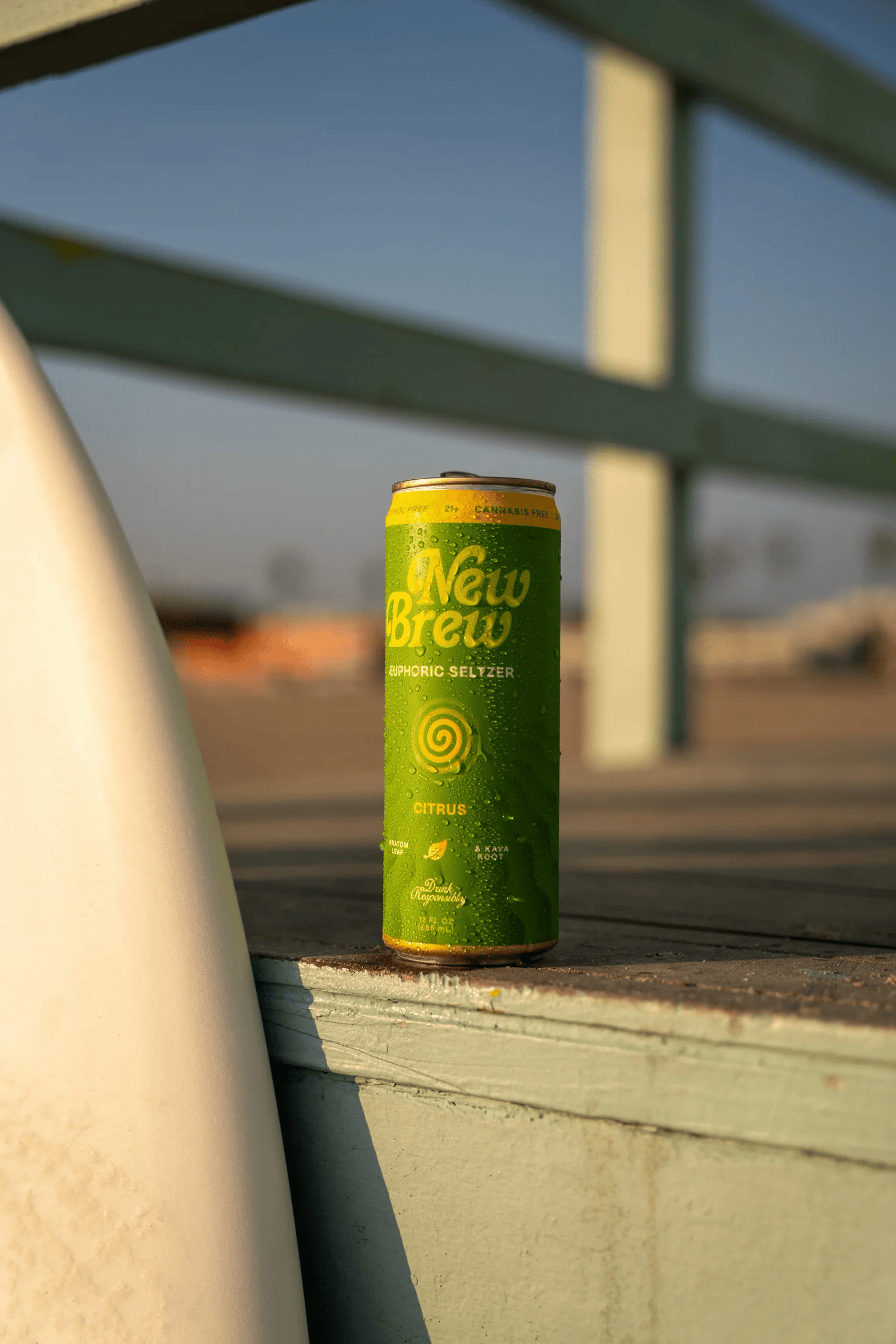 Full-color product photograph of a canned beverage balanced on a boardwalk railing at golden hour