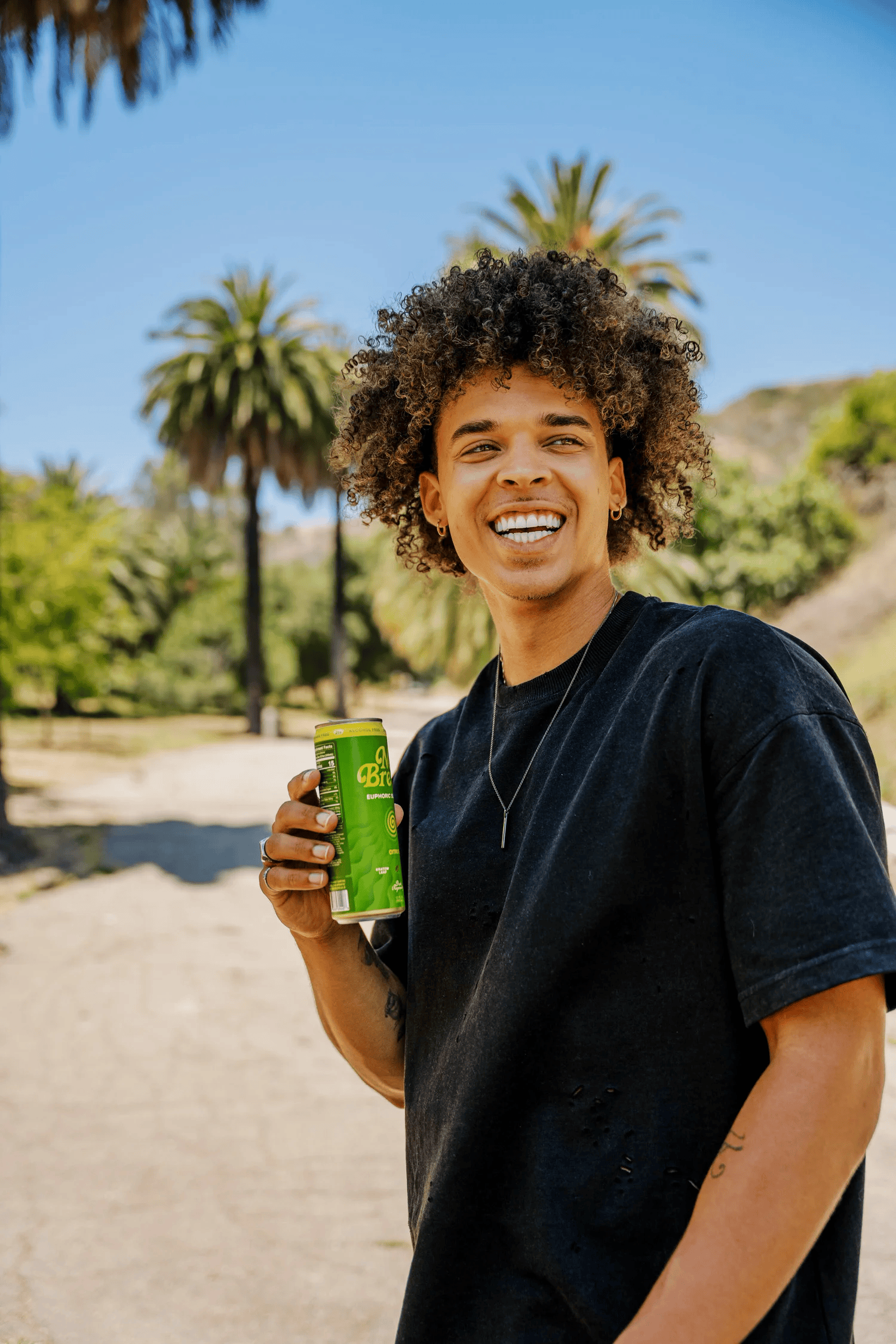 Full-color lifestyle campaign photograph of a smiling man holding a canned beverage on a palm-lined path