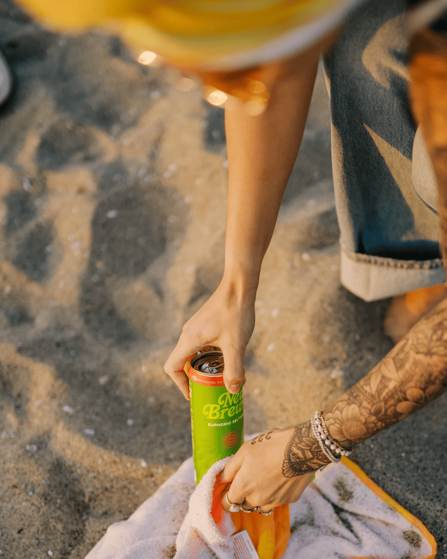 Full-color lifestyle campaign photograph of a canned beverage held over a sandy beach towel at sunset