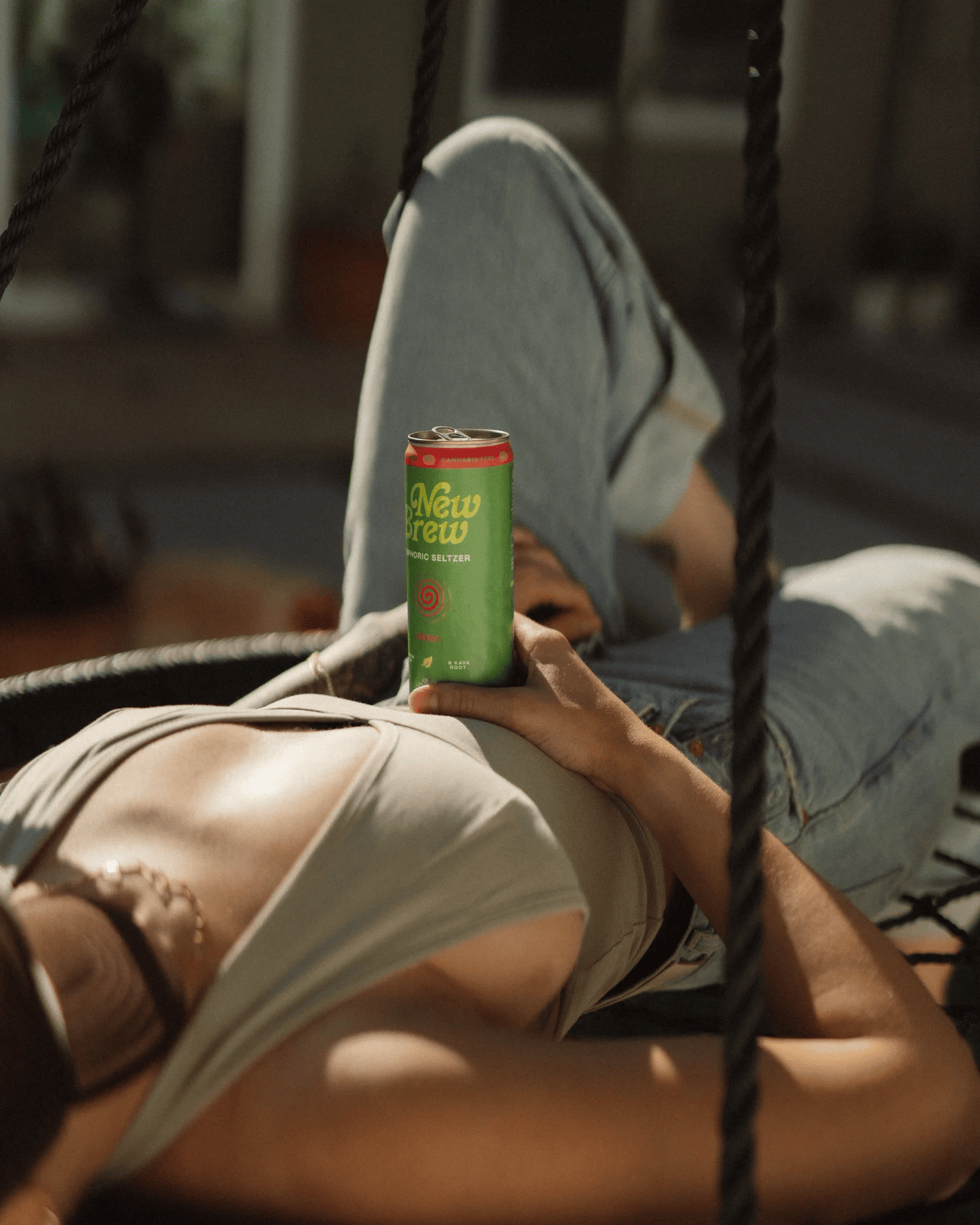 Full-color lifestyle campaign photograph of a canned beverage resting on a woman's lap on an outdoor swing