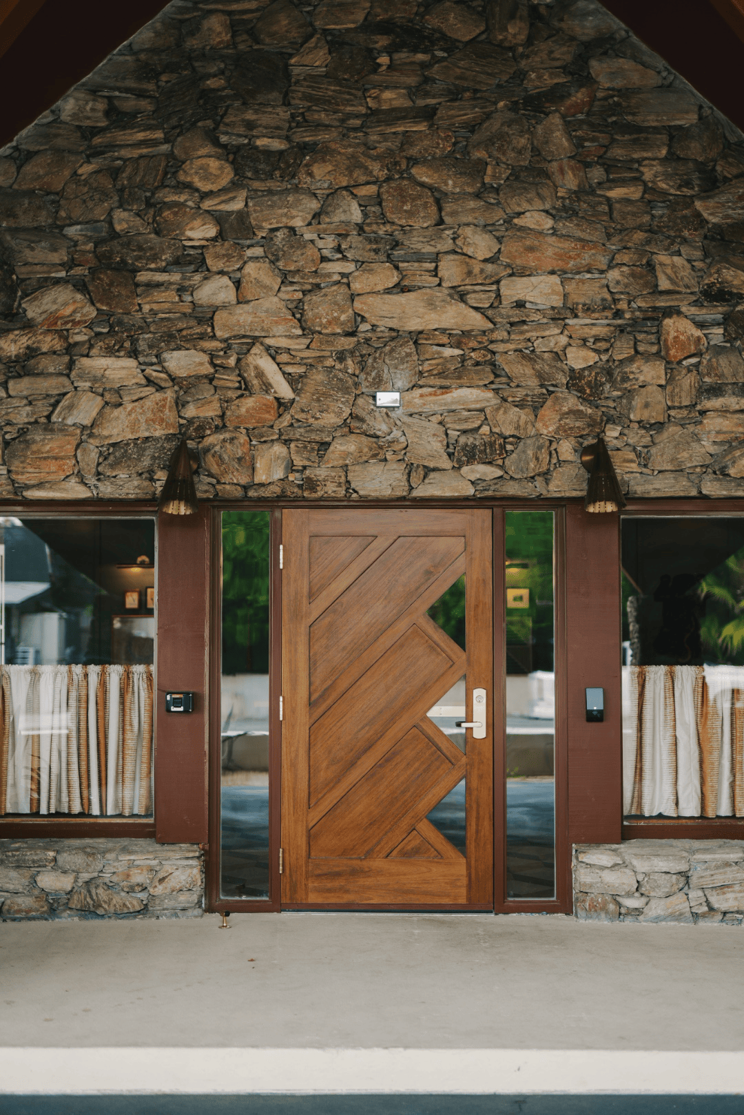 Stone gable facade with a geometric wood front door flanked by sidelight windows and striped curtains beyond, full-color architectural photograph