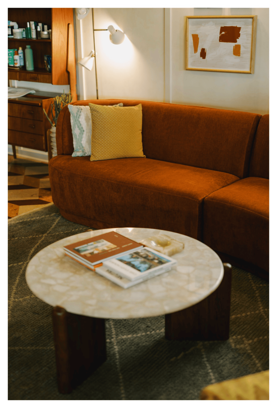 Rust-colored sofa with patterned throw pillows beside a round marble coffee table stacked with books, framed abstract art on the wall behind, full-color interior photograph