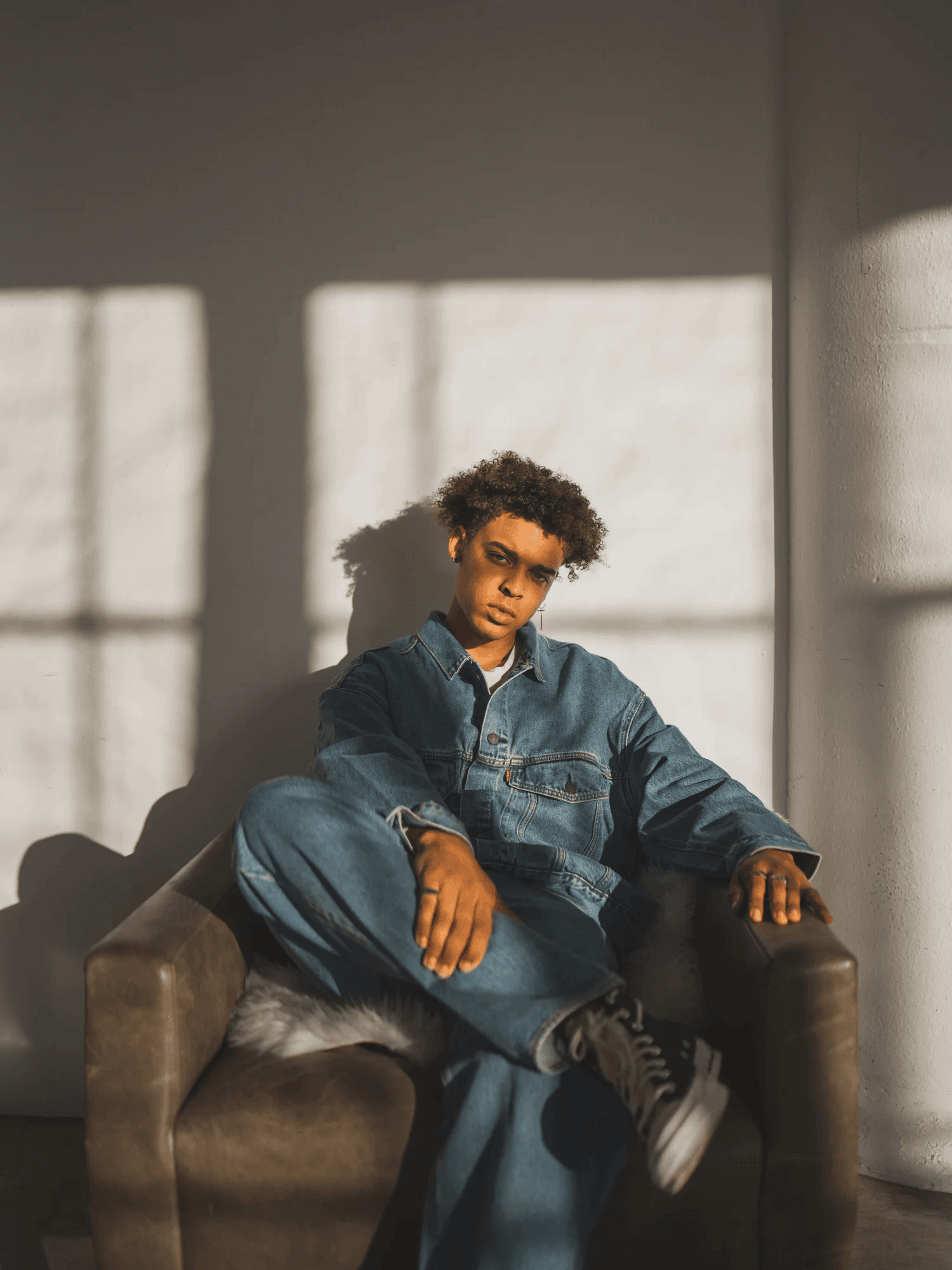 Full-color portrait of a young man in a denim jacket seated in a leather armchair with window-light shadows