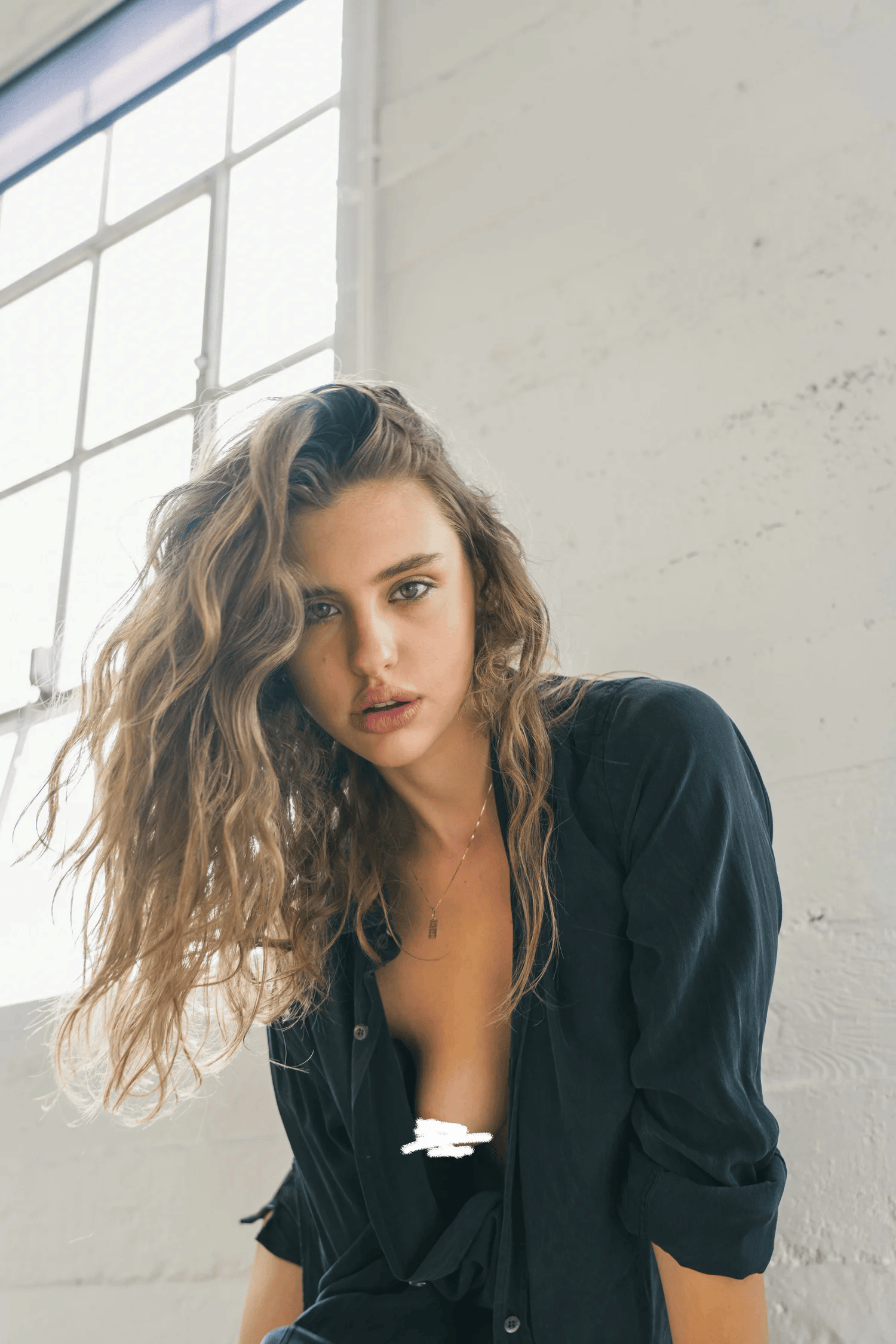 Full-color close-up portrait of a woman with tousled hair in an unbuttoned black shirt against a whitewashed wall