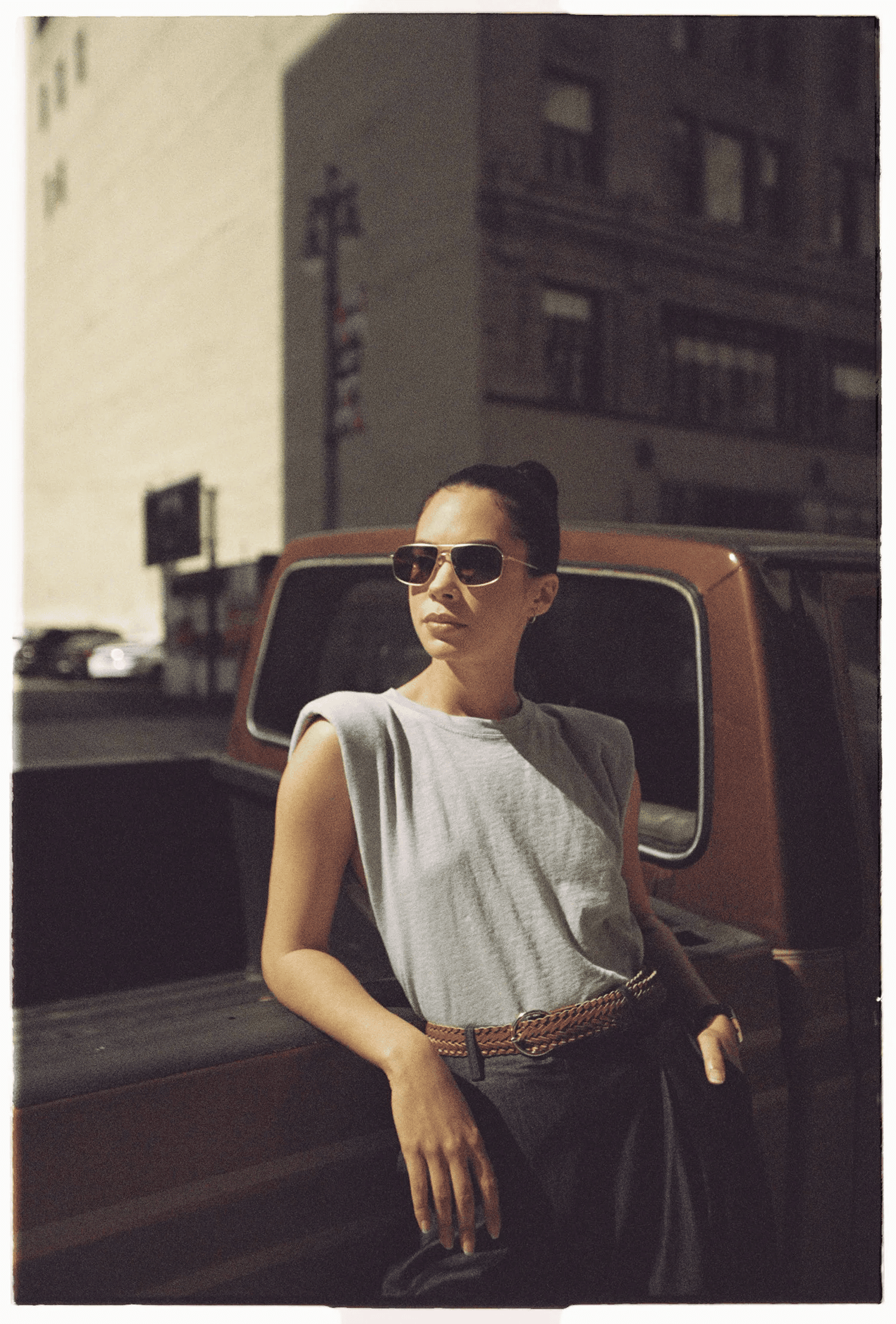 Full-color film-toned street portrait of a woman leaning against a vintage pickup truck in sunglasses